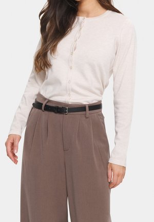 Vrouw draagt een crèmekleurige blouse met lange mouwen en knopen, ingestopt in bruine broek met hoge taille en een zwarte riem.