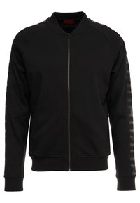 Giacca nera con zip e maniche raglan, polsini a costine e un dettaglio con logo lungo il braccio sinistro. Realizzata in tessuto liscio con una texture morbida.