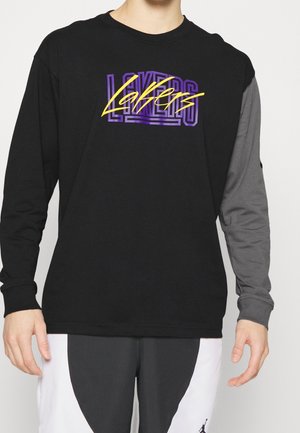Osoba nosząca czarną bluzkę z długim rękawem z fioletowym i żółtym tekstem "Lakers" oraz niedopasowanymi czarnymi i szarymi rękawami.