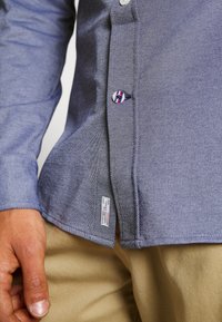 Camicia blu a maniche lunghe con motivo strutturato, bottoni bianchi e un bottone con logo. Etichetta visibile sull'orlo, abbinata a pantaloni tan a contrasto.