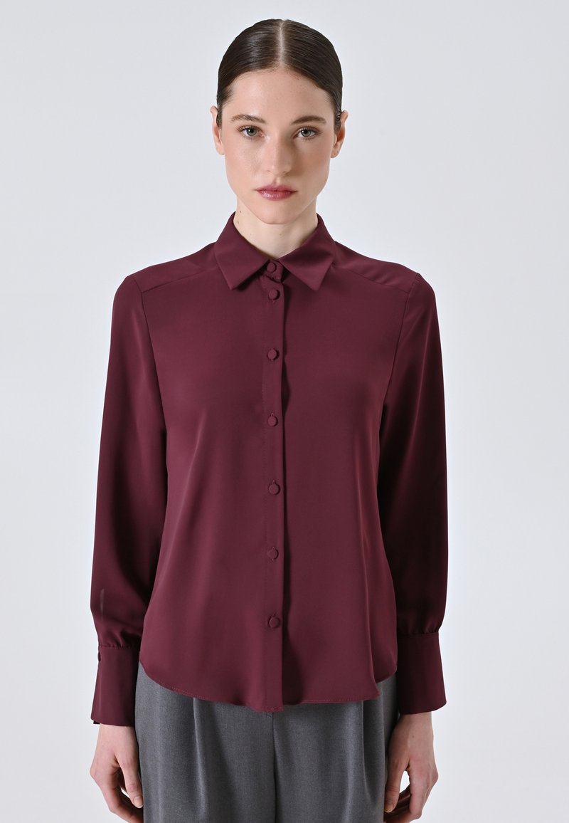 Bordeaux zijden blouse met een knoopsluiting aan de voorkant, spitse kraag en lange mouwen met knoopmanchetten. Zachte textuur en afgeronde zoom.