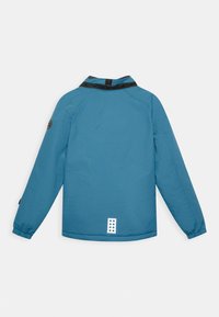 Leichte blaue Jacke mit schwarzem Kragen, elastischen Bündchen und einem Design, das hinten ein weißes rechteckiges Detail mit runden Aussparungen aufweist.