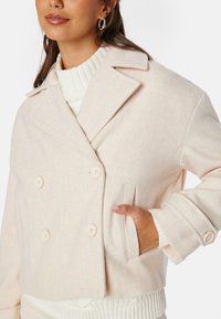 Dvouřadový růžový kabát z vlněné směsi s velkými knoflíky, klopovými límci a bočními kapsami. Pod ním je vidět pružný bílý svetr s límcem.
