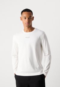 Bărbat tânăr cu părul scurt, îmbrăcat cu un tricou alb cu mâneci lungi pe care scrie "Marc O'Polo" și pantaloni negri, stând cu mâinile în buzunare.