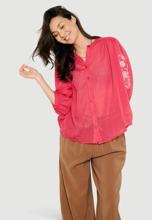 Frau mit dunklen Haaren, die lächelt und eine lockere rote Bluse mit Knöpfen und floraler Stickerei an den Ärmeln sowie braune Hosen mit weitem Bein trägt.