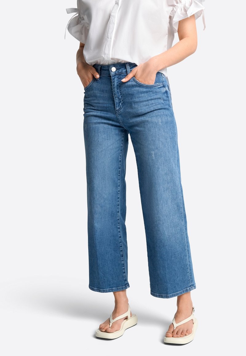 Donna che indossa jeans blu a gamba larga crop, una blusa bianca a maniche corte con volant e sandali platform bianchi, in piedi con le mani nelle tasche.