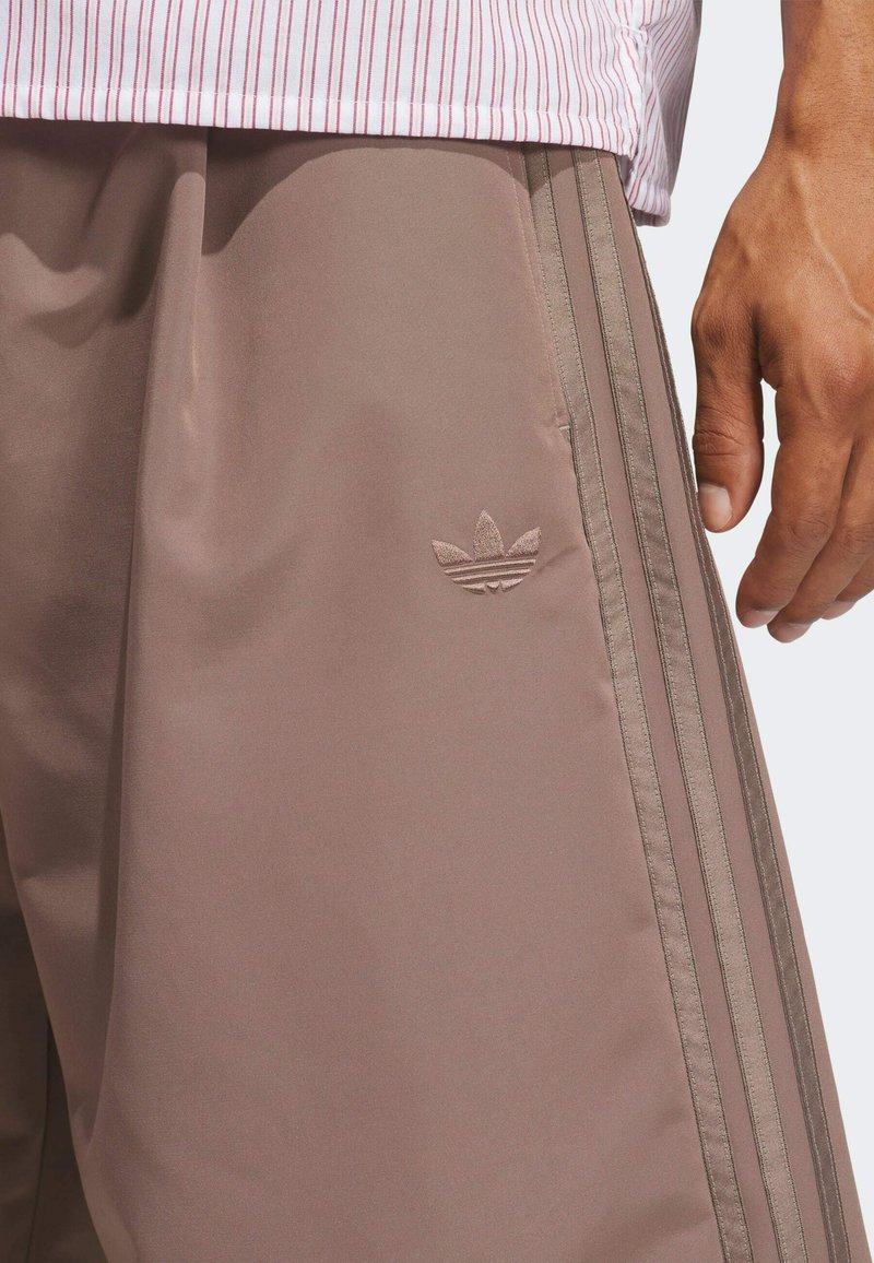 Close-up of person wearing brown Adidas track pants with three side stripes and Trefoil logo, paired with pink and white striped shirt.