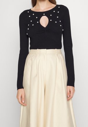 Mujer con top negro de manga larga adornado con perlas y abertura en forma de lágrima, combinado con pantalones beige de cintura alta y pernera ancha.