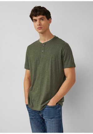Jonge man met kort krullend haar, gekleed in een olijfgroen henley-shirt met korte mouwen en een blauwe spijkerbroek, staande met handen in de zakken.