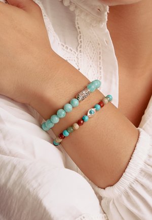 Arm mit zwei Perlenarmbändern, eines mit großen Türkisperlen und silbernem Akzent, das andere mit mehrfarbigen kleinen Perlen und einem silbernen ovalen Anhänger.