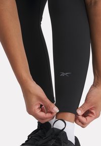 Leggings atletici neri con una texture liscia, vita alta e logo riflettente vicino alla caviglia. Indossati con sneakers nere e calzini bianchi.