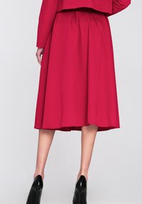 Czerwona spódnica do kolan wykonana z gładkiego materiału, z plisami w pasie i rozkloszowanym krojem. Pod spodem widoczne czarne szpilki.