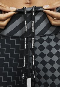 Czarne legginsy sportowe z geometrycznym wzorem, z wiązaniem do regulacji z napisem "NIKE RUNNING". Zbliżenie na pas.