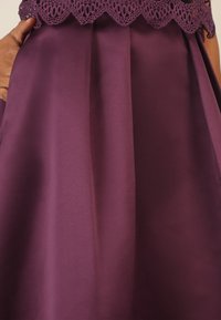 Abito in satin viola con dettaglio in pizzo scallopato al collo e gonna plissettata, che mette in risalto la superficie liscia e il design elegante.