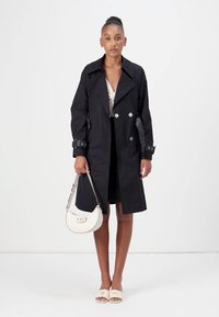 Zwarte trenchcoat met dubbele knopen, zijzakken en verstelbare manchetten, gecombineerd met een witte handtas en lichte schoenen.