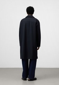 Cappotto lungo nero con una vestibilità ampia, caratterizzato da una silhouette dritta, design minimal e una trama liscia. Pantaloni in denim larghi.
