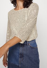 Maglione beige con paillettes, maniche a tre quarti, scollatura rotonda e design a maglia testurizzata, abbinato a una gonna in denim.