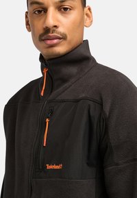 Giacca in fleece grigio scuro con una tasca con zip nera, tirazip arancione e logo Timberland sul davanti, caratterizzata da un design con colletto alto.