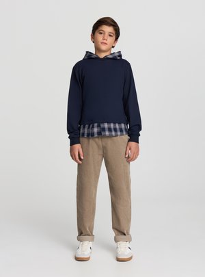 Granatowa bluza z kapturem nałożona na niebieską koszulę w kratę, zestawiona z beżowymi spodniami sztruksowymi i białymi trampkami z czarnymi akcentami.