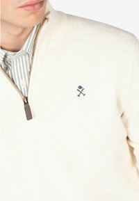 Suéter texturizado color crema con un diseño de cremallera de cuarto, tirador de cremallera beige y un logo bordado en azul marino en el lado izquierdo del pecho.