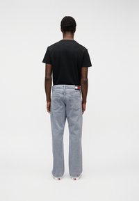 Άντρας που στέκεται με την πλάτη στην κάμερα, φοράει μαύρο μπλουζάκι, ανοιχτόχρωμα μπλε τζιν και λευκά παπούτσια, μπροστά σε απλό λευκό φόντο.