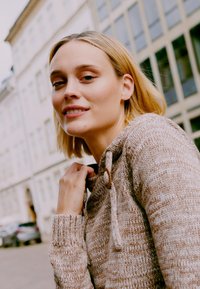 Beige strikket genser med et teksturert design, med hette og lukking med treknapper. Modellen trekker forsiktig i kragen.