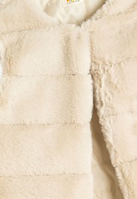 Beige Kunstfellweste mit strukturiertem Oberflächenmaterial, ausgestattet mit horizontalen Nähten und einem weichen Innenfutter. Offene Vorderseite, leicht.