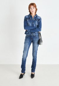 Veste en denim et jeans ajustés de couleur bleu moyen, avec des coutures classiques, cinq poches et un sac à main noir. Le mannequin porte des chaussures à talons noires.