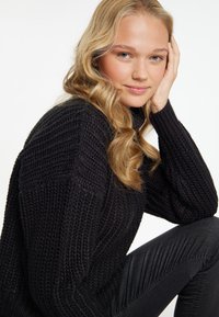 Schwarzer Strickpullover mit einem strukturierten Rippenmuster, hohem Kragen und langen Ärmeln. Getragen mit dunklen Skinny-Jeans.