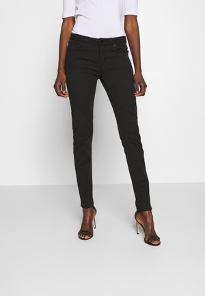 Vrouw met slanke zwarte broek en zwarte sandalen met hoge hakken die tegen een lichte achtergrond staat.