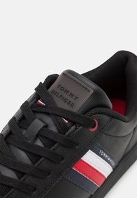 Gros plan d'une basket noire Tommy Hilfiger montrant les lacets, la languette en cuir avec le logo de la marque, et des rayures rouges, blanches et bleues sur le côté.