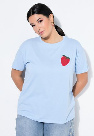 Vrouw met donker haar in een paardenstaart, die een lichtblauw T-shirt draagt met een rode aardbei-afbeelding op de borst en lichtblauwe spijkerbroek.