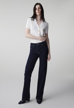 Donna in piedi che indossa una maglietta bianca a maniche corte con bottoni e jeans scuri a vita alta e gamba dritta, abbinati a scarpe nere appuntite.