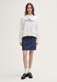 Overdimensjonert kremfarget sweatshirt med marineblå "TOM TAILOR" tekst, kombinert med et tettsittende mørkt denimskjørt og svarte plattformloafers. Hvite ankelstrømper.