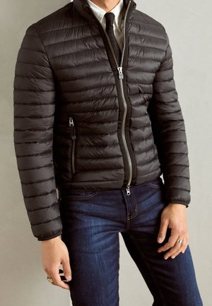 Homme portant une veste matelassée noire à fermeture éclair sur une chemise blanche et une cravate noire, associée à un jean bleu foncé, debout contre un mur uni.