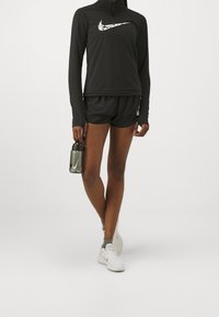 Idrottare i svart långärmad tröja och shorts med vita sneakers, håller en mörk vattenflaska med en sportvarumärkeslogotyp.