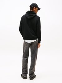 Junger Mann steht seitlich, trägt einen schwarzen Hoodie, dunkelgraue Jeans, schwarze Sneakers und eine schwarze Kappe, vor einem weißen Hintergrund.