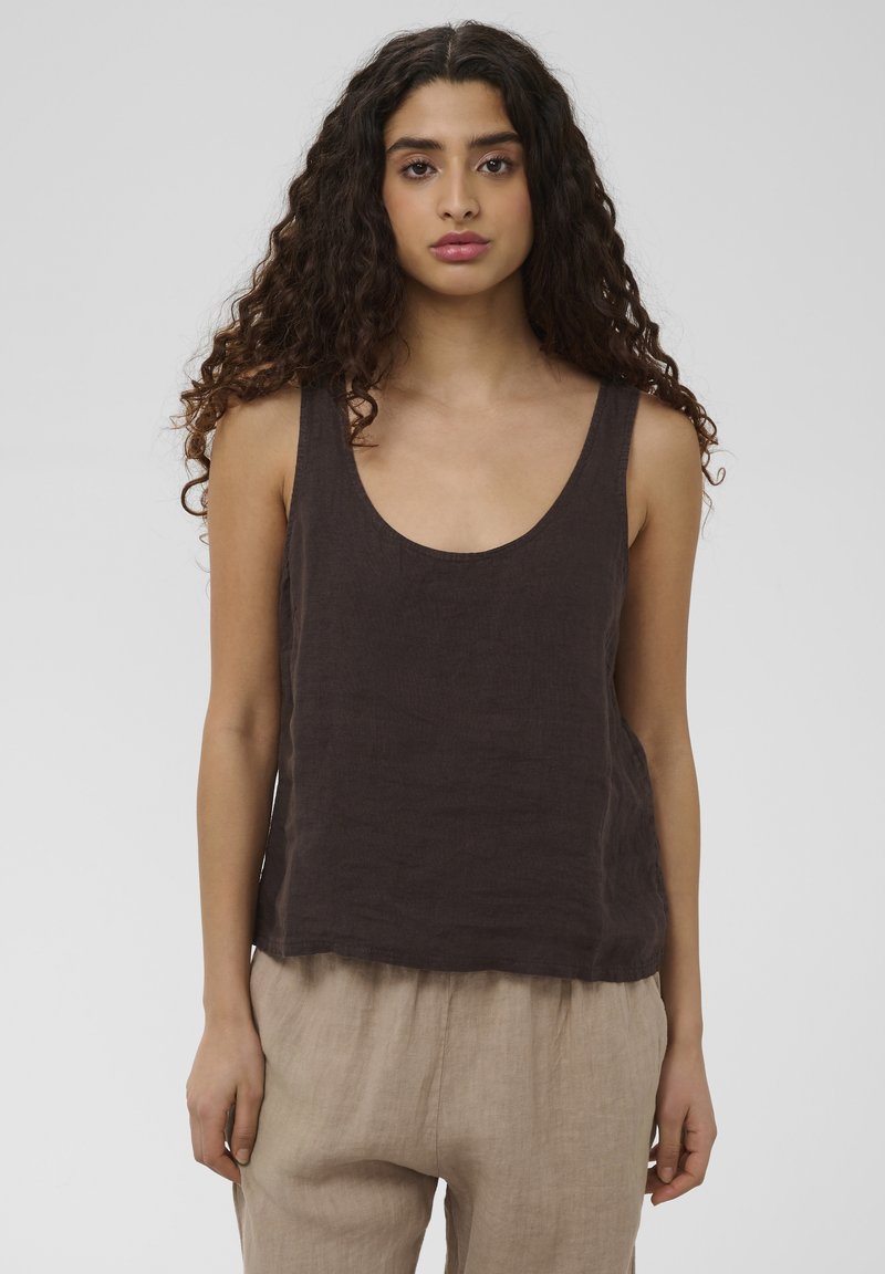 Jeune femme aux longs cheveux bouclés portant un haut sans manches foncé et un pantalon beige sur un fond clair uni.