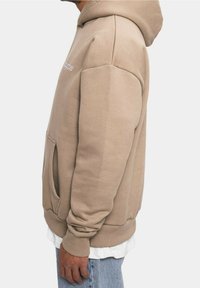 Sudadera con capucha beige hecha de tela suave, con un bolsillo tipo canguro, puños y dobladillo acanalados. El diseño es de corte holgado y cuenta con un logo visible.