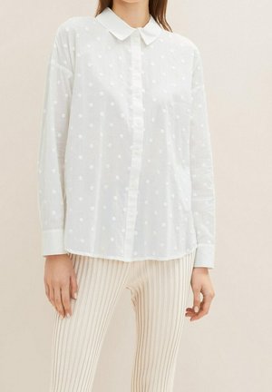 Femme portant une chemise blanche à manches longues boutonnée avec une texture à pois et un pantalon côtelé beige.