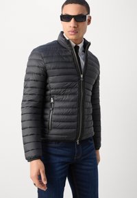 Doudoune noire avec un matelassage horizontal, un col montant et une fermeture éclair à l'avant. Elle possède deux poches latérales zippées et une texture lisse.