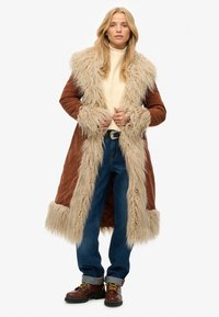 Brun ruskindsfrakke med en bred krave og manchetter i kunstpels. Har et langt design, parret med blå jeans og brune snørebåndsstøvler.