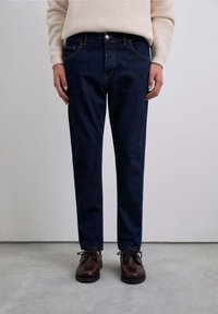 Jeans de mezclilla azul oscuro con un corte ajustado, diseño de cinco bolsillos y costuras amarillas. Combinados con zapatos de cuero marrón.