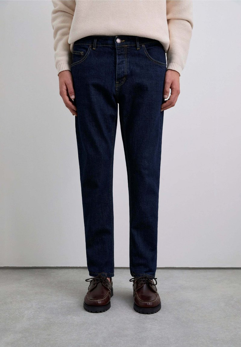 Jeans din denim albastru închis, cu o croială slim, design cu cinci buzunare și cusături galbene. Asociate cu pantofi din piele maro.
