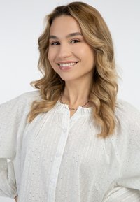 Fehér, texturált blúz gombos elülső résszel, apró ezüst részletekkel és ráncolt nyakkivágással. A modellen hullámos haj van, és egy egyszerű nyakláncot visel.