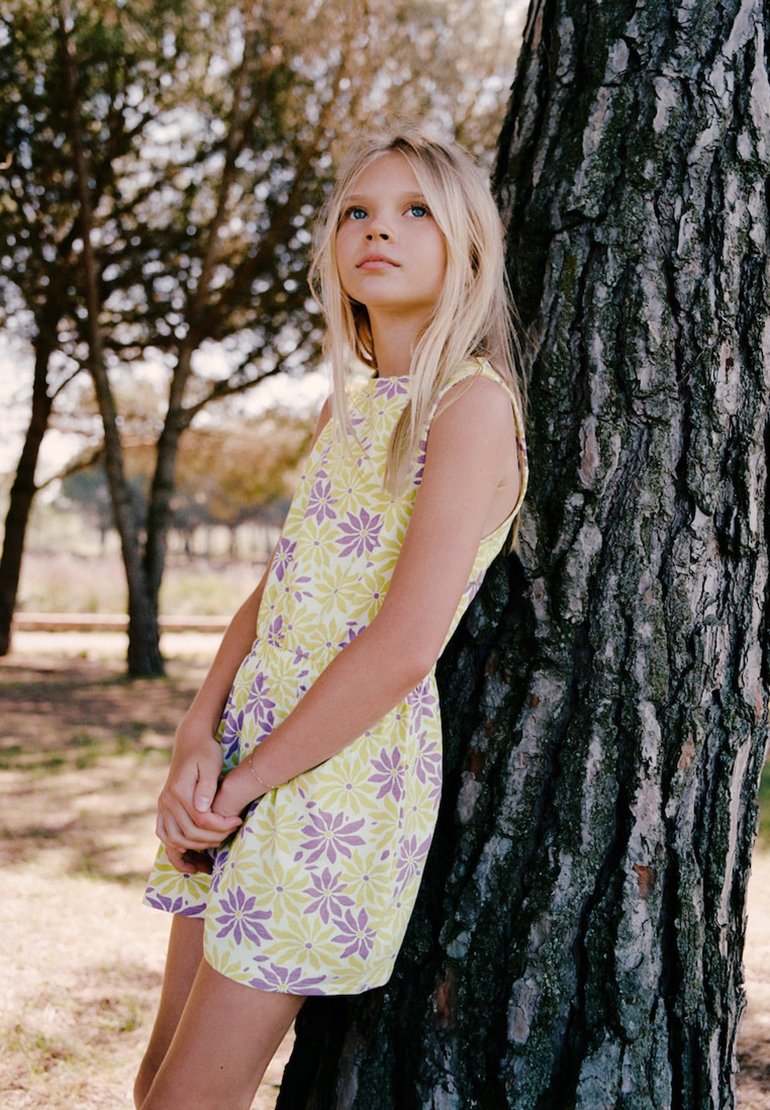 Jeune fille aux longs cheveux blonds portant une robe fleurie jaune et violette, appuyée contre un arbre en plein air sous une lumière douce.