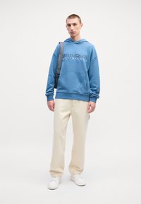 Hoodie di cotone blu con logo "HUGO", tasca frontale, abbinato a pantaloni beige e sneakers bianche, il modello porta anche una borsa a spalla grigia.