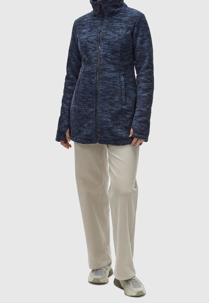 Personne portant une veste zippée bleu marine avec poches latérales, pantalon beige à larges jambes et baskets grises et blanches, debout devant un fond uni.