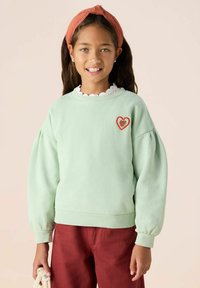Sweatshirt vert clair avec des manches bouffantes, des poignets côtelés et une broderie de cœur rouge sur la poitrine. Porté avec un pantalon brun-rouge.