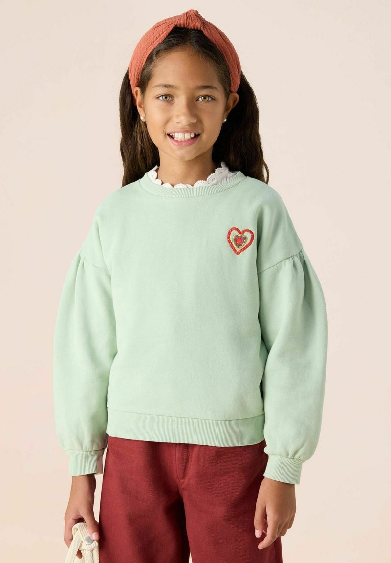 Sweatshirt vert clair avec des manches bouffantes, des poignets côtelés et une broderie de cœur rouge sur la poitrine. Porté avec un pantalon brun-rouge.